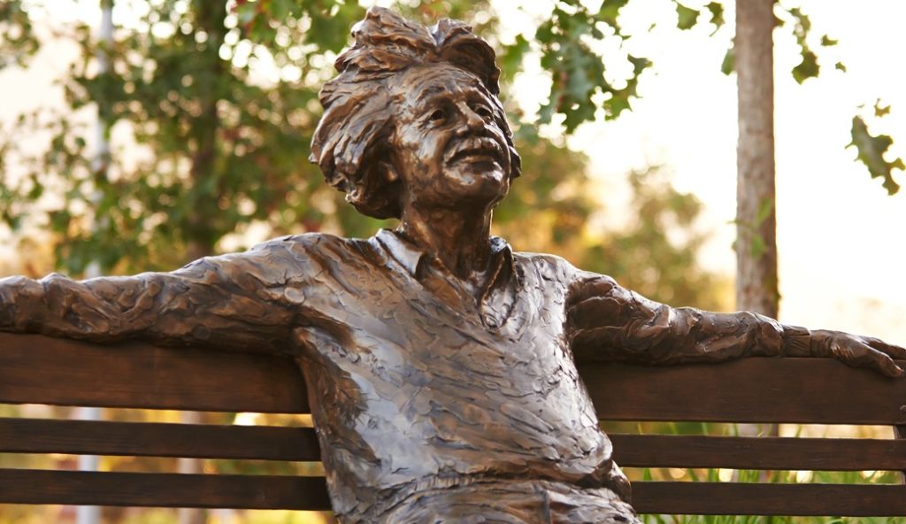 Bronze scientist statue with outstretched arms sitting on a park bench