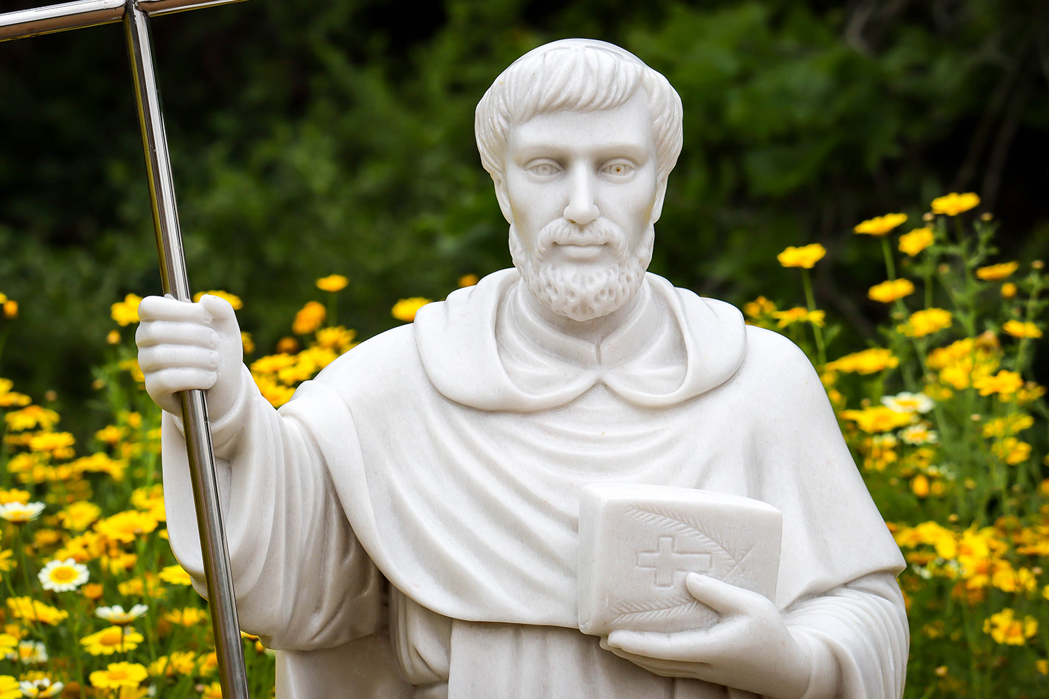A white statue of a saint holding a cross and a book in a flower garden